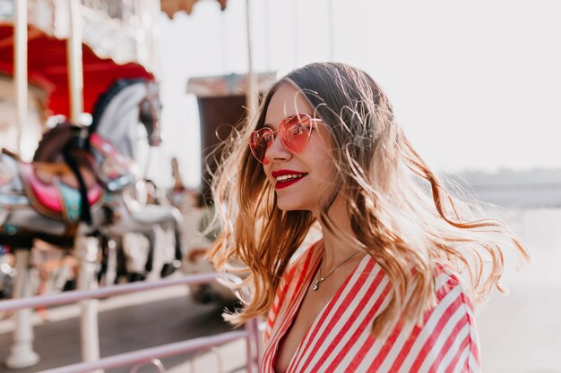 Menina bonita andando em um parque de diversões. Modelo feminino sorridente em óculos de sol cor de rosa da moda tem um bom dia no verão.