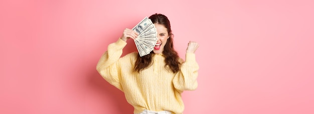 Menina bonita animada dizendo sim ganhar dinheiro do prêmio segurando notas de dólar na metade do rosto fazendo o punho