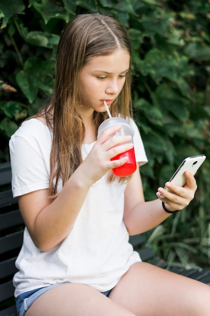 Menina bonita bebendo suco enquanto estiver usando smartphone no parque