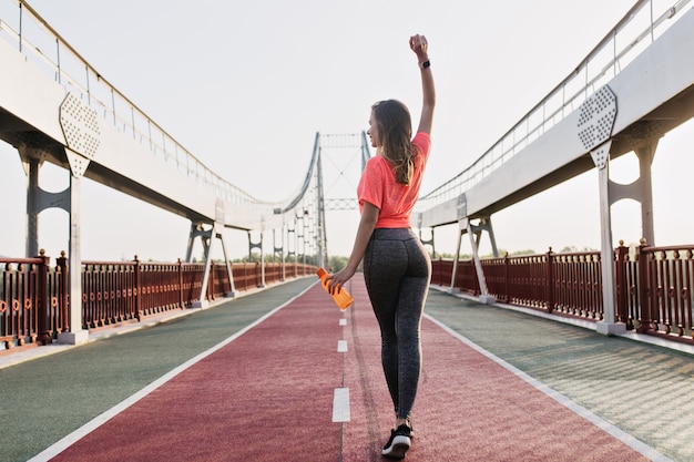 Foto grátis menina bonita branca estendendo-se no estádio de manhã cedo. foto ao ar livre da parte de trás de uma mulher refinada fazendo exercícios.