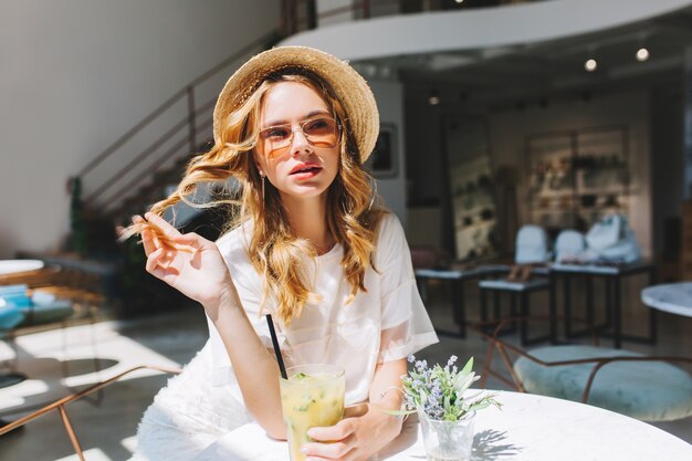 Menina bonita brincando com seu cabelo encaracolado descansando na mesa com um pequeno vaso de flores sobre ele