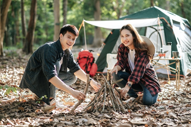 Menina bonita campista preparando lenha com o namorado para iniciar uma fogueira. Jovem casal de turistas ajudando a colher galhos e montá-los na frente de uma barraca de acampamento