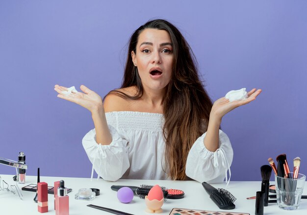 Menina bonita chocada sentada à mesa com ferramentas de maquiagem segurando mousse de cabelo isolada na parede roxa