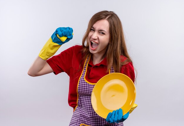 Menina bonita com avental e luvas de borracha, lavando o prato com a esponja, gritando com expressão agressiva