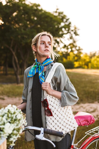 Menina bonita com bicicleta e bolsa branca sonhadoramente olhando de lado passando tempo no parque