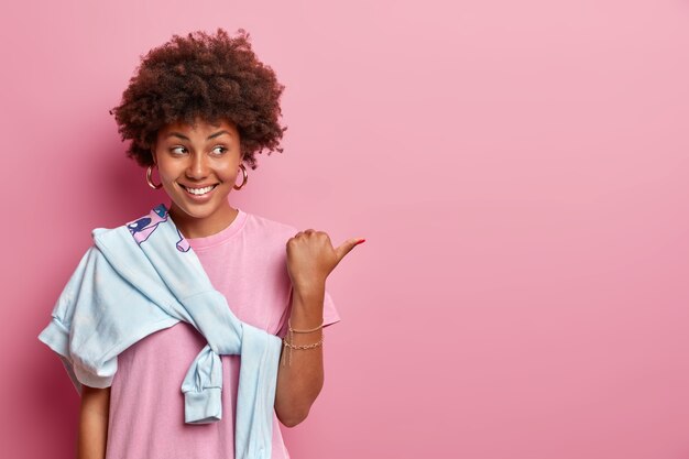 Menina bonita com cabelo afro ajuda a escolher a melhor escolha, aponta o polegar para o lado no espaço da cópia, anuncia o produto, sorri feliz, veste camiseta rosada e suéter amarrado no ombro. Sua promoção aqui