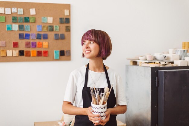 Menina bonita com cabelo colorido no avental preto e camiseta branca segurando a caneca com ferramentas de cerâmica nas mãos sonhadoramente olhando de lado no estúdio de cerâmica