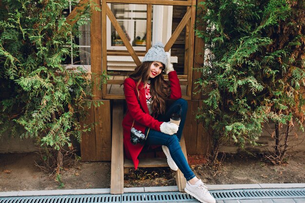 Menina bonita com cabelo comprido, casaco vermelho e chapéu de malha, sentado na escada de madeira ao ar livre. Ela segura a câmera e o café em luvas brancas, sorrindo.