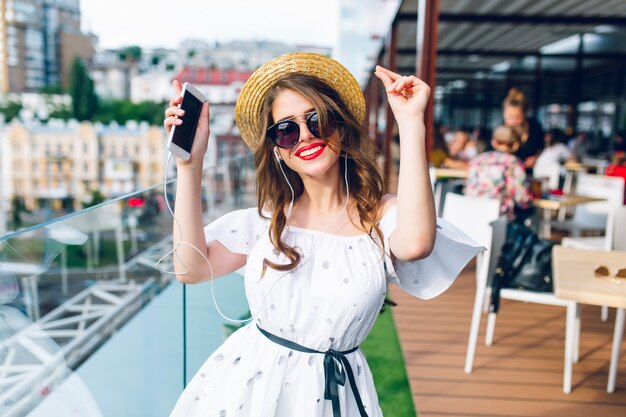 Menina bonita com cabelo comprido em óculos de sol está ouvindo música através de fones de ouvido no terraço. Ela usa um vestido branco com ombros nus, batom vermelho e chapéu. Ela está dançando.