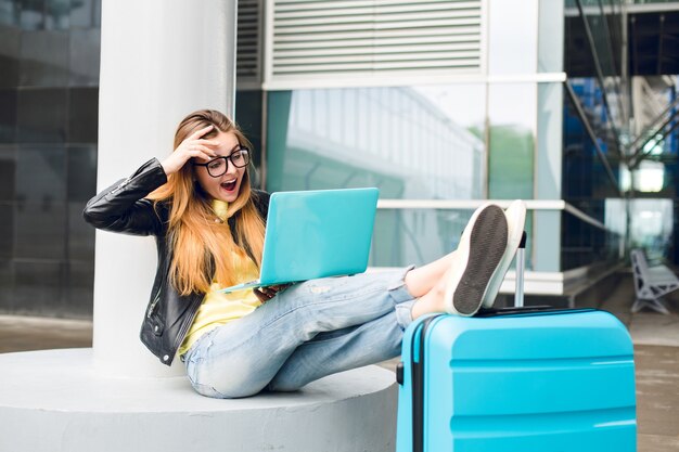 Menina bonita com cabelo comprido em óculos escuros está sentada do lado de fora no aeroporto. Ela usa jeans, jaqueta preta, sapatos amarelos. Ela colocou as pernas na mala e falando no laptop. Ela parece surpresa.