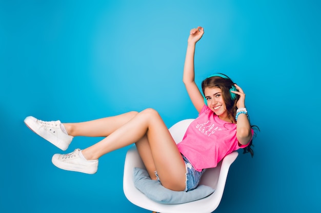 Menina bonita com cabelo longo cacheado, ouvindo música na cadeira sobre fundo azul no estúdio. Ela usa shorts, camiseta rosa, tênis branco. Ela segura as pernas acima e sorri para a câmera.