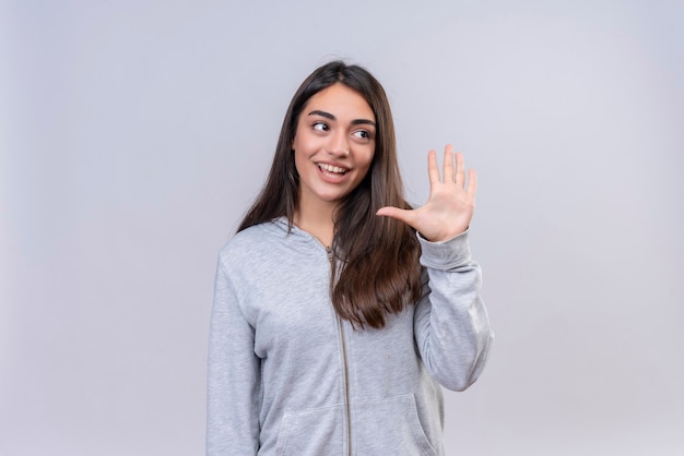 Menina bonita com capuz cinza olhando para longe, mostrando cinco com um sorriso no rosto, de pé sobre um fundo branco