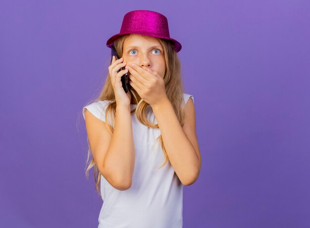 Menina bonita com chapéu de férias falando no celular, ficando chocada, conceito de festa de aniversário em pé sobre fundo roxo