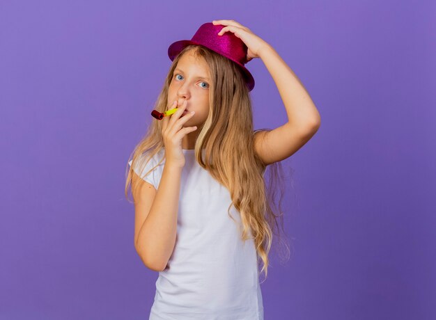 Menina bonita com chapéu de férias soprando apito feliz e positivo, conceito de festa de aniversário em pé sobre fundo roxo