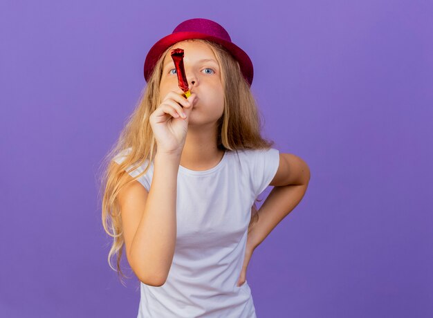 Menina bonita com chapéu de férias soprando apito feliz e positivo, conceito de festa de aniversário em pé sobre fundo roxo