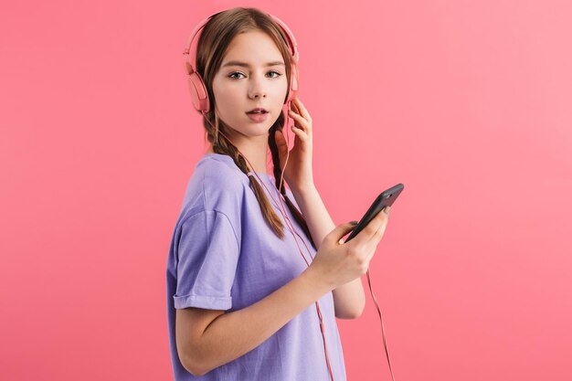 Menina bonita com duas tranças na camiseta lilás com fones de ouvido segurando o celular na mão enquanto sonhadoramente olhando na câmera sobre fundo rosa