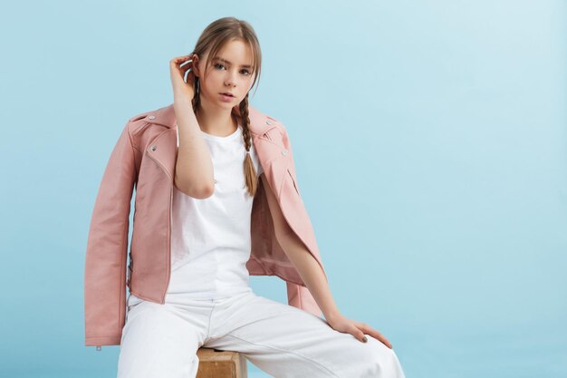 Menina bonita com duas tranças na jaqueta de couro rosa e jeans branco sonhadoramente olhando na câmera enquanto passa o tempo sobre fundo azul