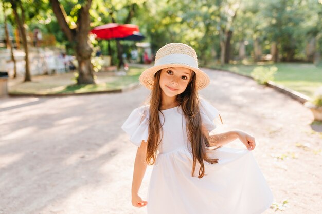 Menina bonita com lindos olhos escuros, posando enquanto se diverte no parque nas férias de verão. Retrato ao ar livre da criança engraçada de cabelos compridos em pé de chapéu de palha na estrada com um sorriso surpreso.