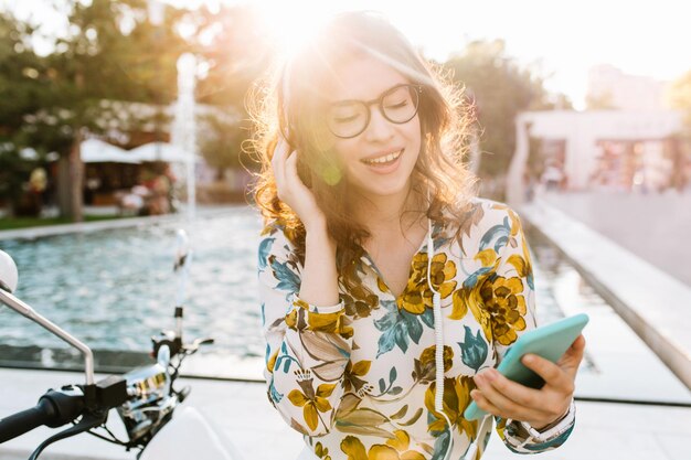 Menina bonita com sorriso surpreso, lendo a mensagem, segurando o telefone e tocando seu cabelo escuro. Retrato ao ar livre de uma jovem deslumbrante em óculos elegantes, descansando ao lado da fonte, depois do trabalho.