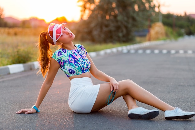 Menina bonita com tampa transparente, top multicolor e saia branca, sentado na estrada ao pôr do sol.