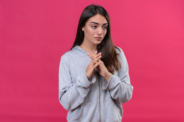 Menina bonita com um casaco cinza com carapuço e uma expressão pensativa em pé sobre um fundo rosa