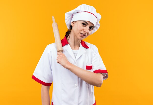 Menina bonita com uniforme de chef satisfeita segurando o rolo de massa no ombro isolado na parede laranja