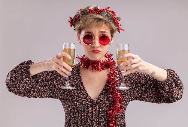 Menina bonita confusa com coroa de flores de natal e guirlanda de ouropel em volta do pescoço com óculos segurando duas taças de champanhe, olhando para a câmera, isolada no fundo branco