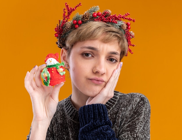 Menina bonita confusa com coroa de flores de natal segurando uma pequena estátua de boneco de neve de natal, mantendo a mão no rosto isolada na parede laranja