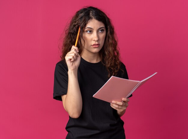 Menina bonita confusa segurando um lápis e um caderno, fazendo um gesto de pensar olhando para o lado