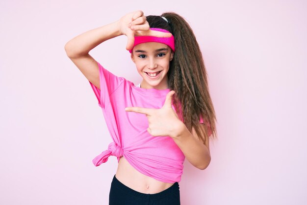 Menina bonita criança hispânica vestindo sportswear sorrindo fazendo moldura com as mãos e os dedos com cara de feliz. conceito de criatividade e fotografia.