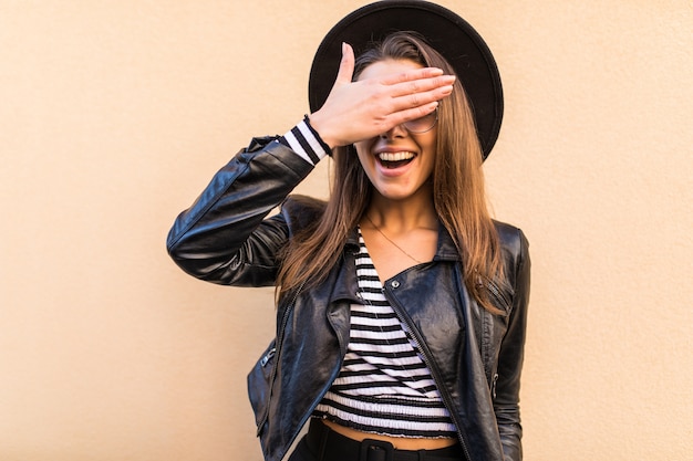 Menina bonita da moda em jaqueta de couro e chapéu preto cobrindo os olhos do rosto com a mão isolada na parede amarela clara