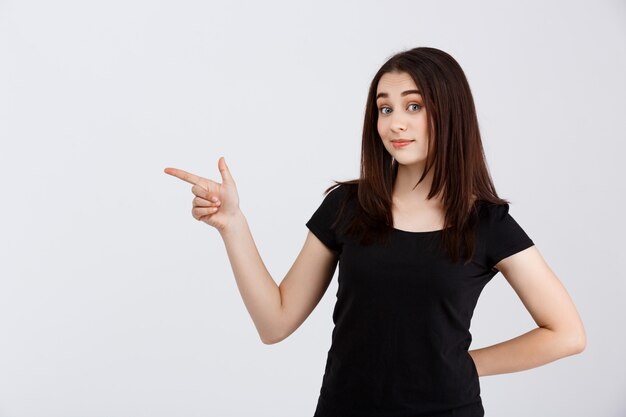 Menina bonita de camiseta preta, apontando os dedos no lado sobre parede branca