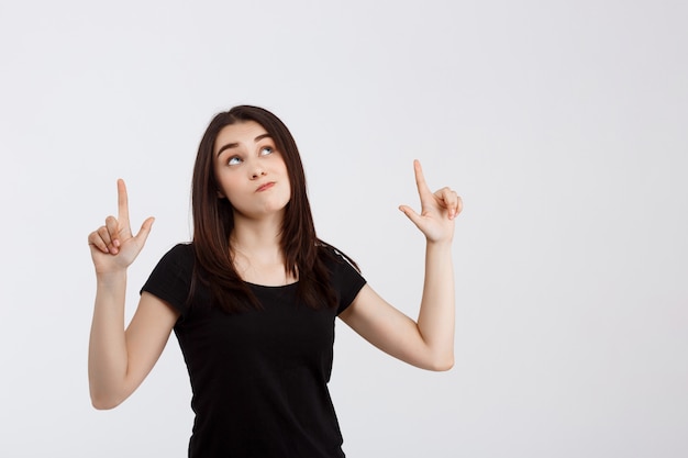 Menina bonita de camiseta preta, apontando os dedos para cima sobre parede branca