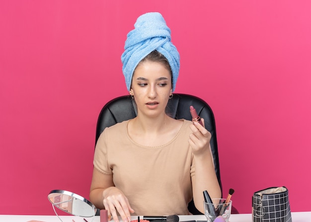 Menina bonita desagradável sentada à mesa com ferramentas de maquiagem enroladas em uma toalha segurando e olhando para o batom isolado no fundo rosa