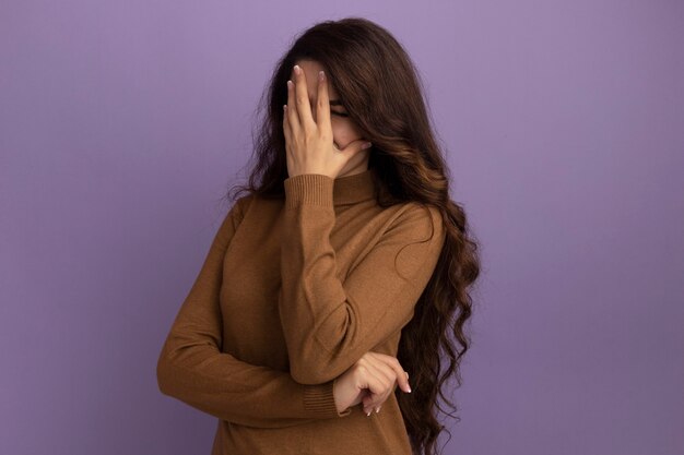 Menina bonita desagradável vestindo um suéter de gola alta marrom coberto com a mão isolada na parede roxa