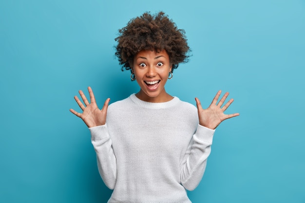 Menina bonita divertida com cabelos afro encaracolados, levanta as palmas das mãos, tem expressão alegre, sorri amplamente, vê algo engraçado, usa suéter branco,