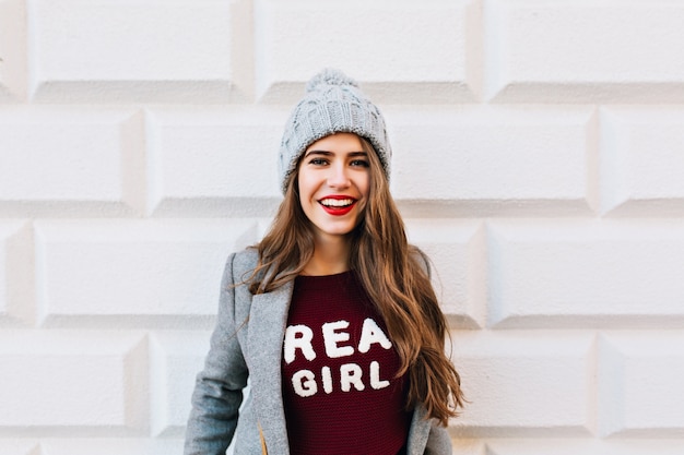 Menina bonita do retrato com cabelo comprido e lábios vermelhos, sorrindo na parede cinza.