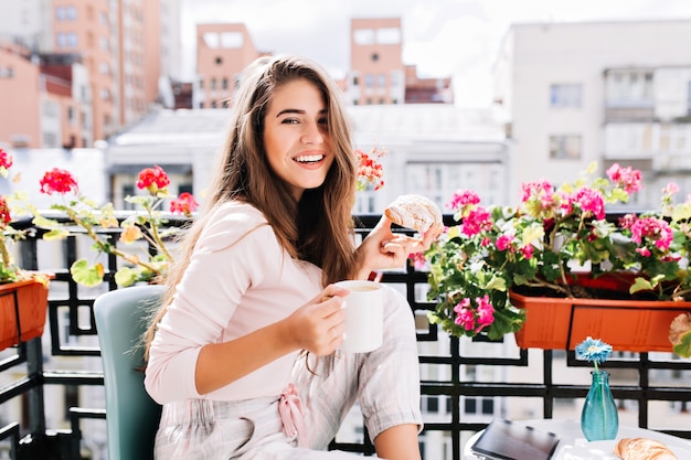 Menina bonita do retrato tomando café da manhã na varanda cercam flores na manhã ensolarada na cidade. Ela segura uma xícara, um croissant, sorrindo.