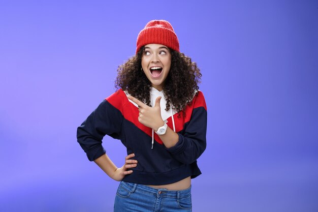 Menina bonita e brincalhona com roupa elegante de inverno e relógio sorrindo amplamente com entusiasmo e alegria apontando, olhando para o canto superior esquerdo emocionado e alegre ao posar sobre um fundo azul.