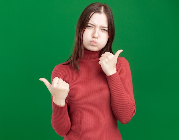 Menina bonita e carrancuda apontando para os lados com bochechas inchadas, isoladas na parede verde com espaço de cópia