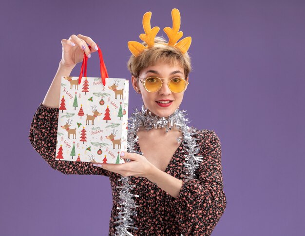 Menina bonita e jovem satisfeita com uma faixa de chifres de rena e uma guirlanda de ouropel no pescoço, com óculos segurando uma sacola de presente de Natal, olhando para a câmera isolada em um fundo roxo com espaço de cópia