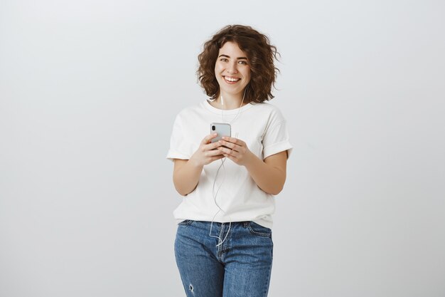 Menina bonita e sorridente ouvindo música em fones de ouvido e trocando mensagens por telefone celular