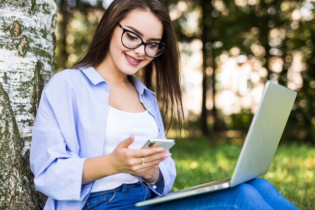 Menina bonita em jeans azul trabalha com laptop e telefone no citypark