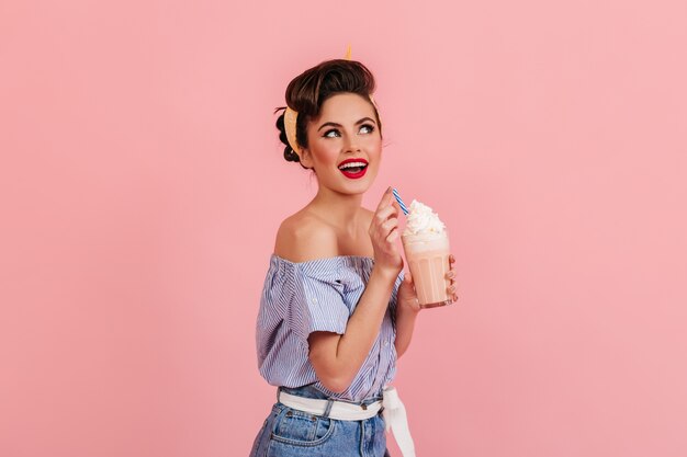Menina bonita espantada posando com milkshake. Mulher jovem na moda desfrutando de um coquetel no fundo rosa.