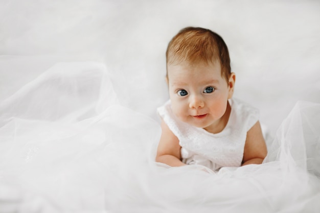 Menina bonita está deitada na barriga com grandes olhos azuis abertos, vestida com roupa branca