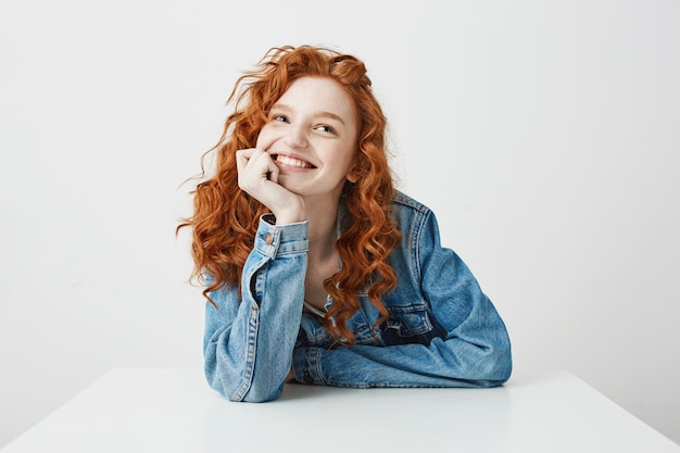 Menina bonita feliz com cabelos cacheados e sardas sorrindo sonhando sentado à mesa.