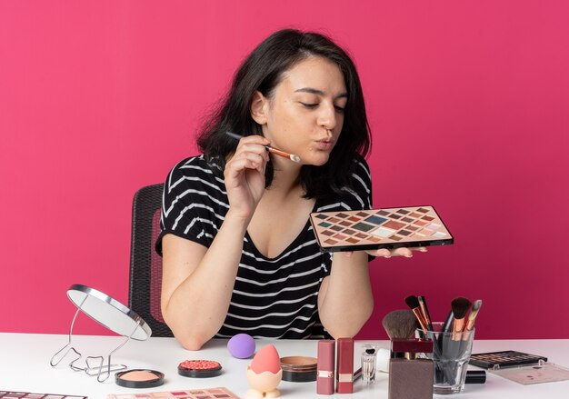 Menina bonita feliz sentada à mesa com ferramentas de maquiagem segurando e olhando para a paleta de sombra com pincel de maquiagem isolado na parede rosa