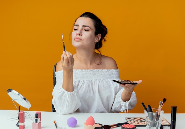 Foto grátis menina bonita irritada se senta à mesa com ferramentas de maquiagem olha para o pincel de maquiagem segurando a paleta de sombras isolada na parede laranja