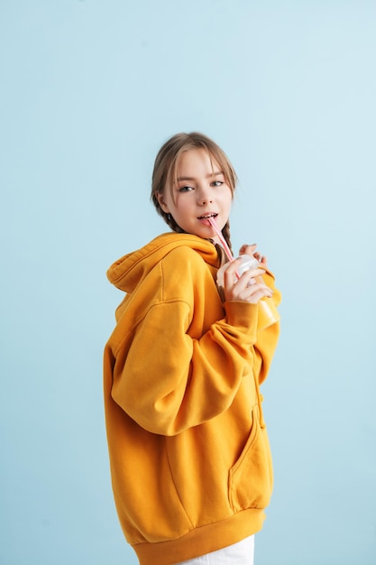 Menina bonita jovem com duas tranças com capuz laranja segurando o copo de plástico com suco na mão enquanto sonhadoramente olhando na câmera sobre fundo azul