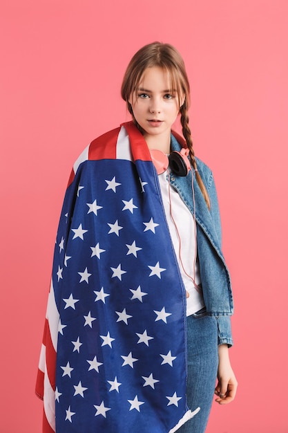Menina bonita jovem com duas tranças na jaqueta jeans com fones de ouvido e grande bandeira americana no ombro sonhadoramente olhando na câmera sobre fundo rosa isolado